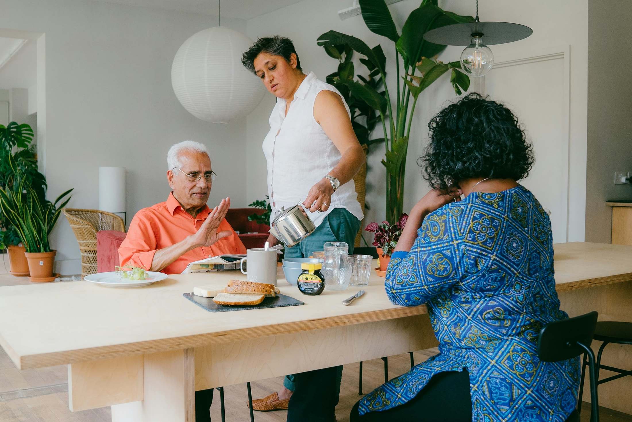 A woman serves breakfast to an older man at a kitchen table at home.